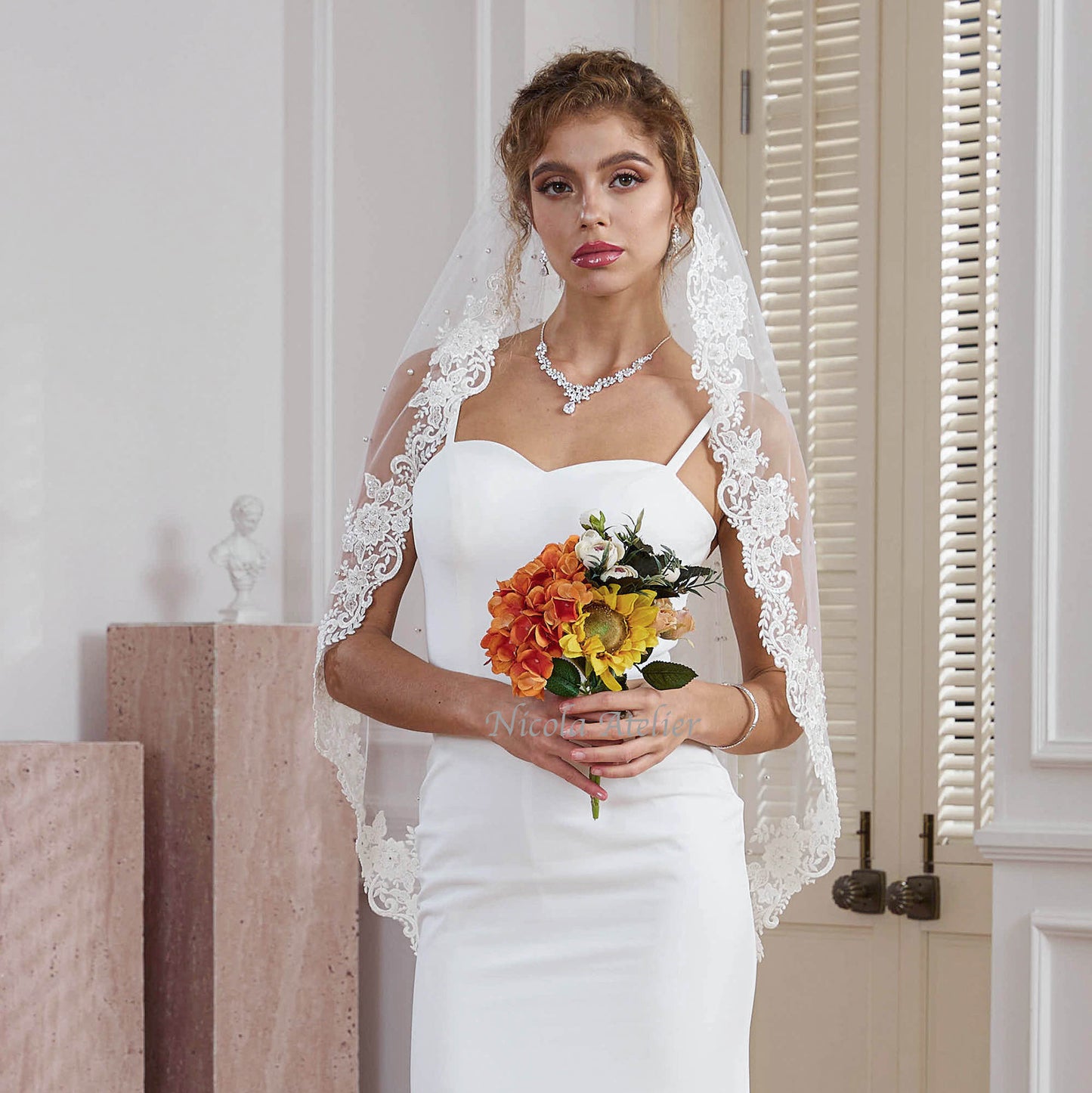 Bride wearing pearl and sequin lace fingertip veil holding warm floral bouquet near bright doorway Nicola Xu Atelier